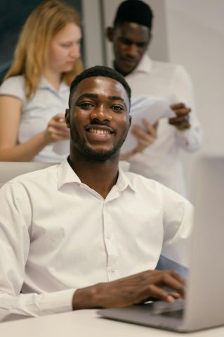 Smiling businessman at desk with colleagues in background. Collaborative office setting.